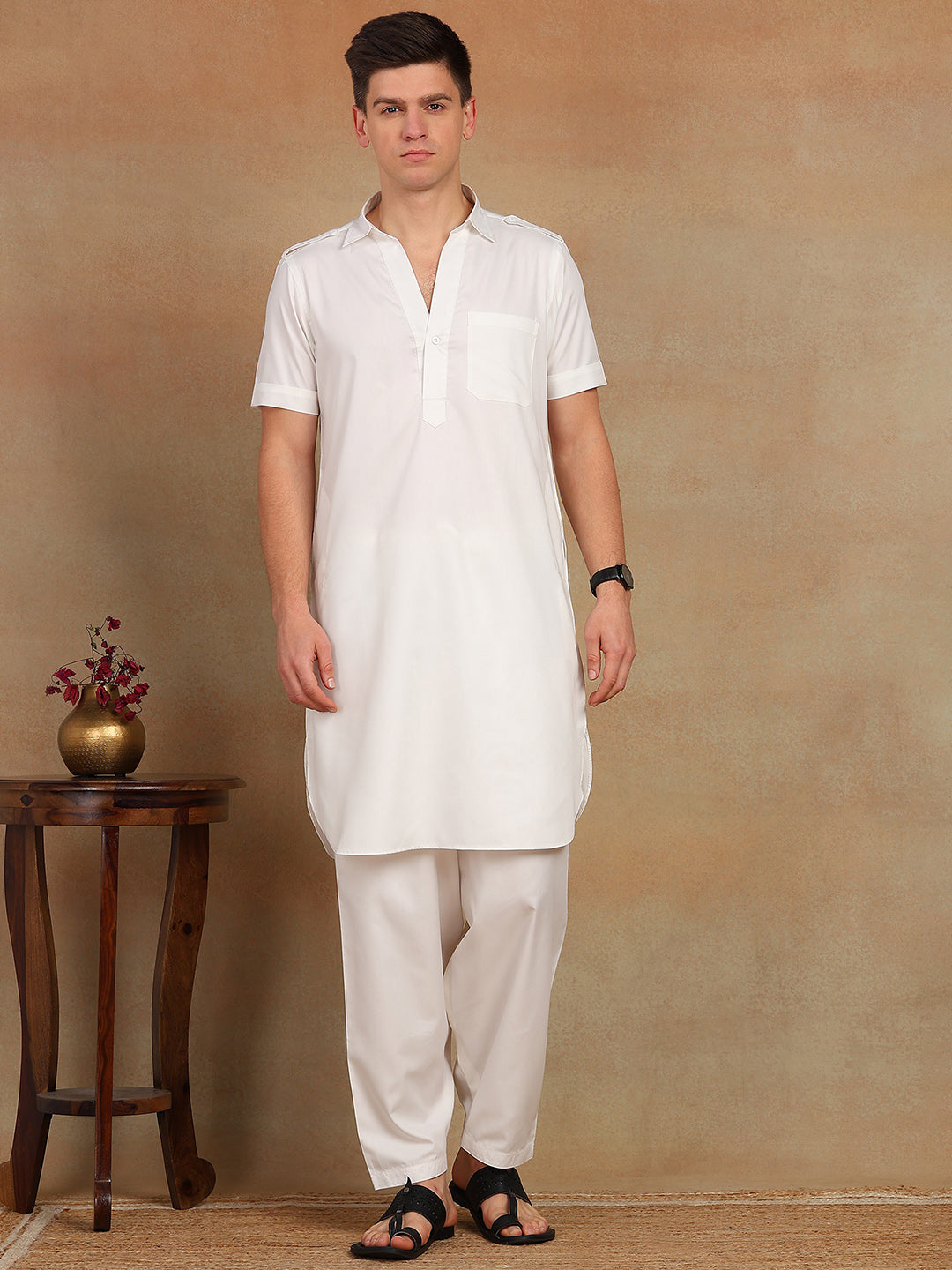 Man wearing a white kurta with white pants against a beige wall.